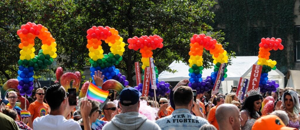 A group of Pride parade-goers holding up rainbow colored balloons that spell out the word Pride